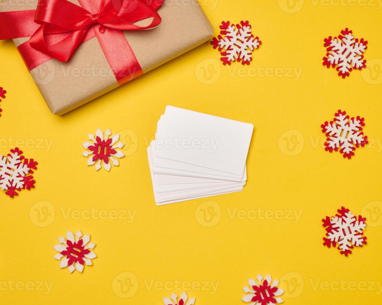 Christmas gift wrapped in red ribbon on yellow background, stack of white paper business cards. Top view photo