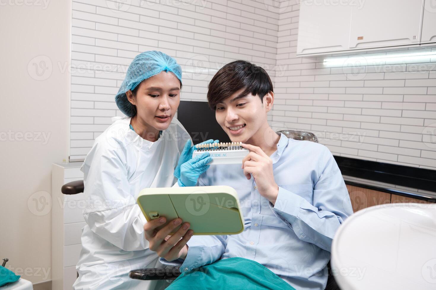 Asian female dentist explains color shades of tooth implant whitening to young male patient, look at mirror in dental clinic, and do professional mouth hygiene healthcare work in a doctor's hospital. photo