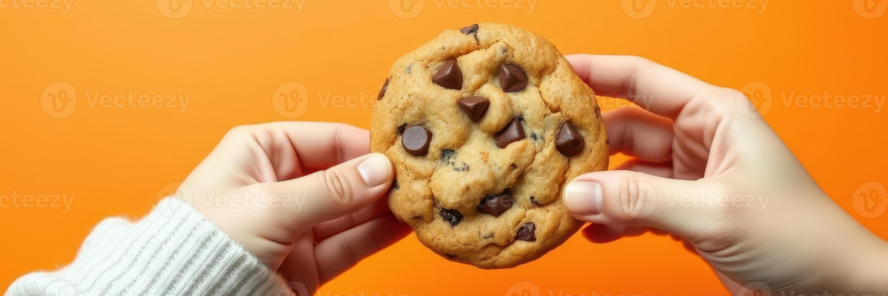 Hands holding a freshly baked chocolate chip cookie against a vibrant orange backdrop photo