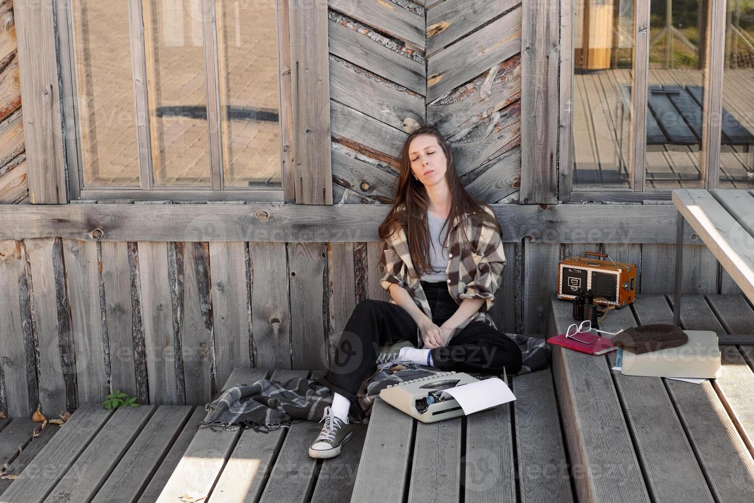 young woman with long hair in a checkered shirt sits thoughtfully on the wooden floor of the terrace next to a typewriter and a retro radio photo