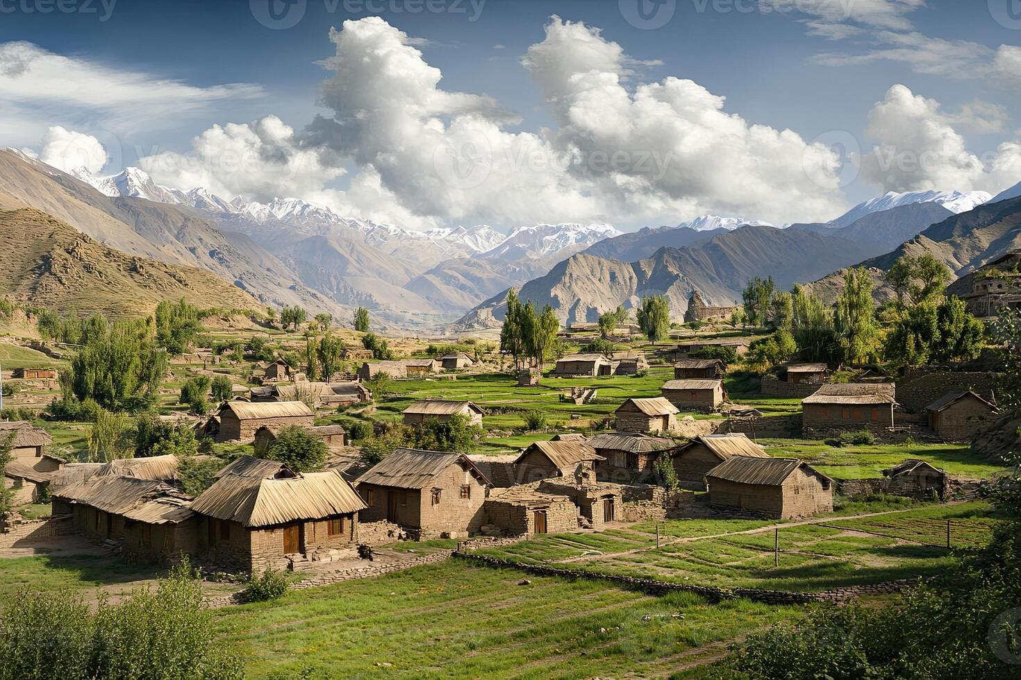 Mountain village nestled in valley with traditional houses, green fields, and snow-capped peaks under a partly cloudy sky. photo