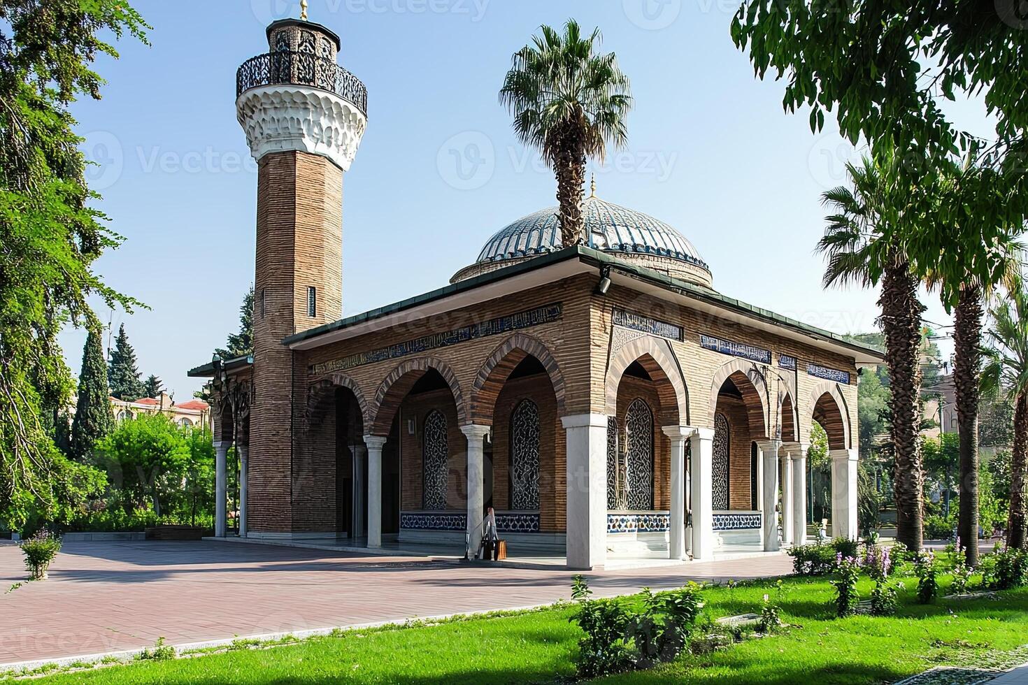 Ottoman-style mosque with a minaret, surrounded by lush greenery and palm trees, under a clear sky. photo