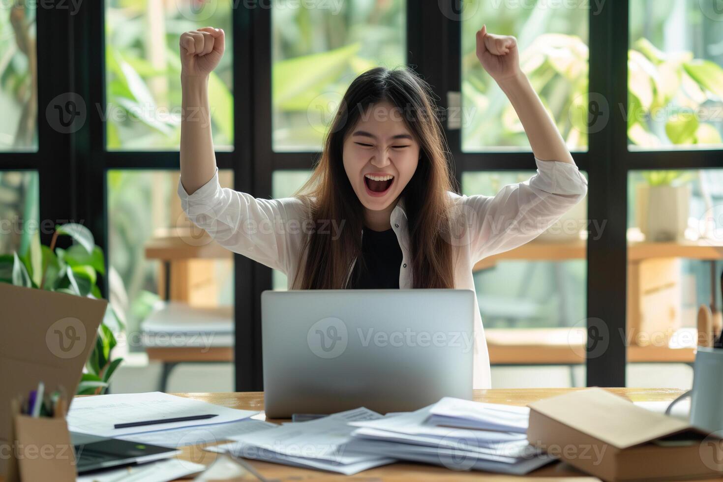 Happy excited young Korean business woman accountant standing at the desk working on laptop computer with a pile of documents on table in office and making yes gesture rejoicing in successful job. photo