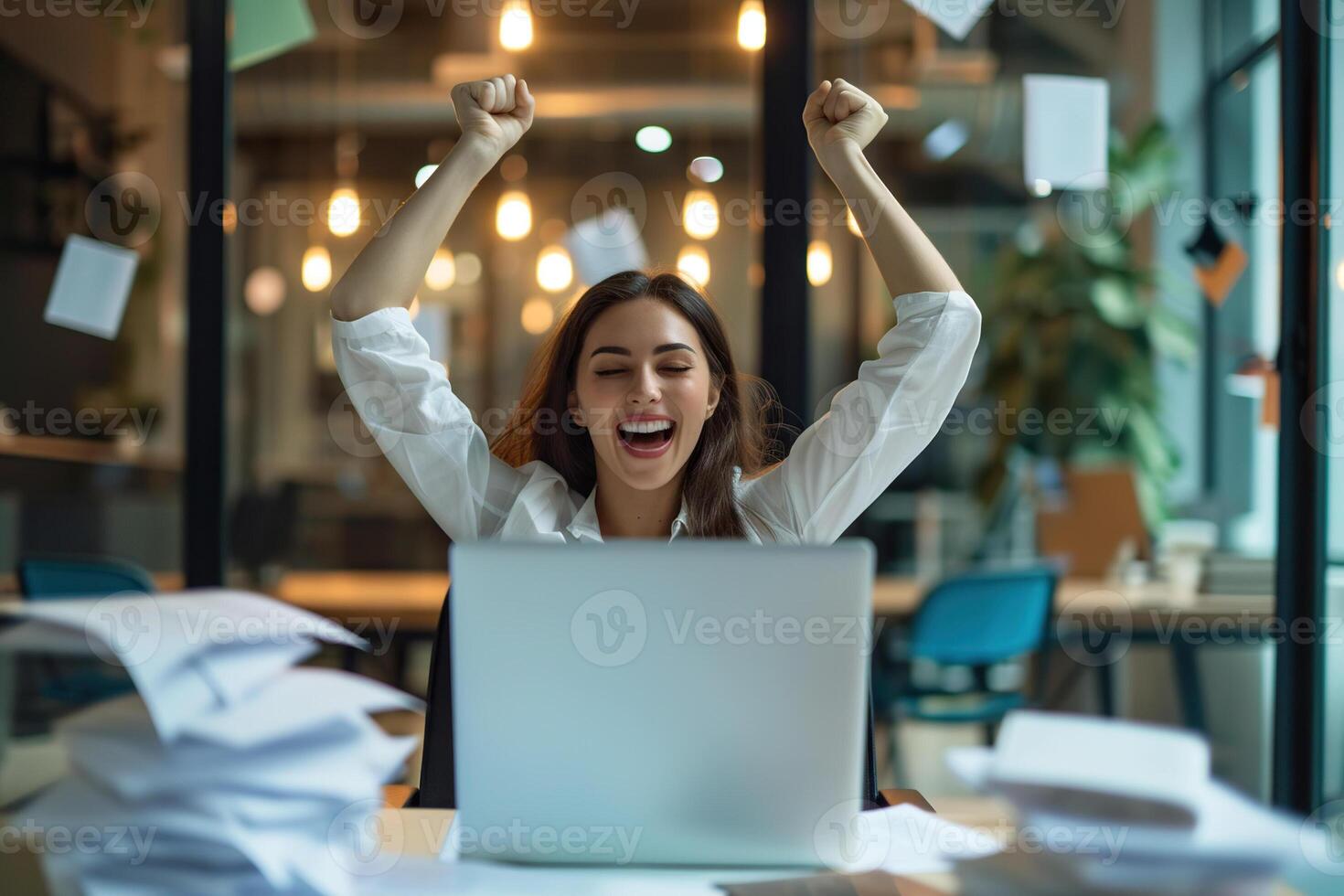 Happy excited young Hispanic business woman accountant standing at the desk working on laptop computer with a pile of documents on table in office and making yes gesture rejoicing in successful job. photo