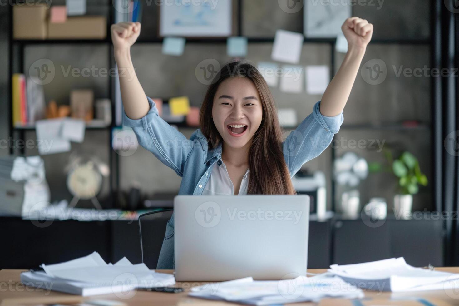 Happy excited young Korean business woman accountant standing at the desk working on laptop computer with a pile of documents on table in office and making yes gesture rejoicing in successful job. photo