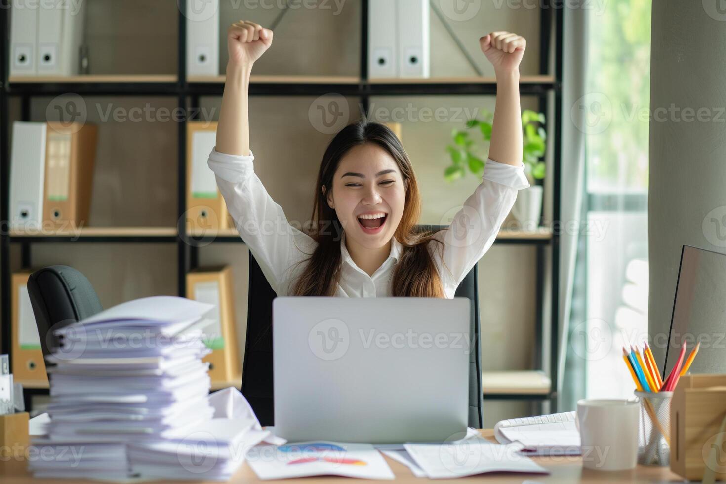 Happy excited young business woman accountant standing at the desk working on laptop computer with a pile of documents on table in office and making yes gesture rejoicing in successful job. photo