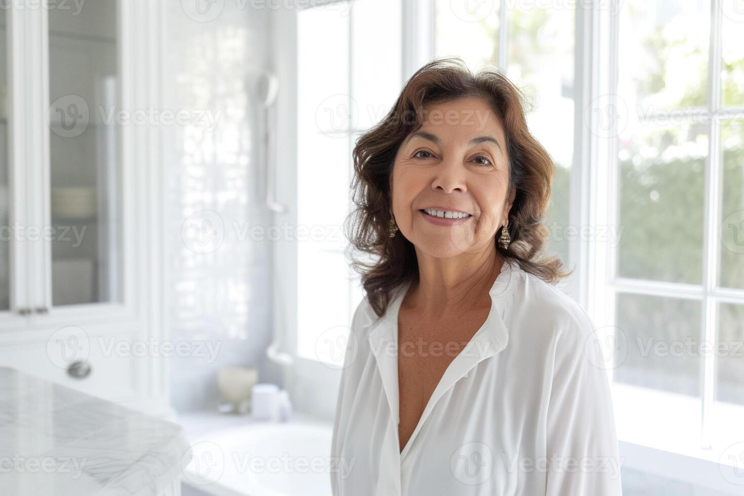 Beautiful, radiant 60 year old Hispanic woman, exuding elegance and vitality with her wide smile, standing in white blouse against the background of a white bathroom and big windows. Copy space photo