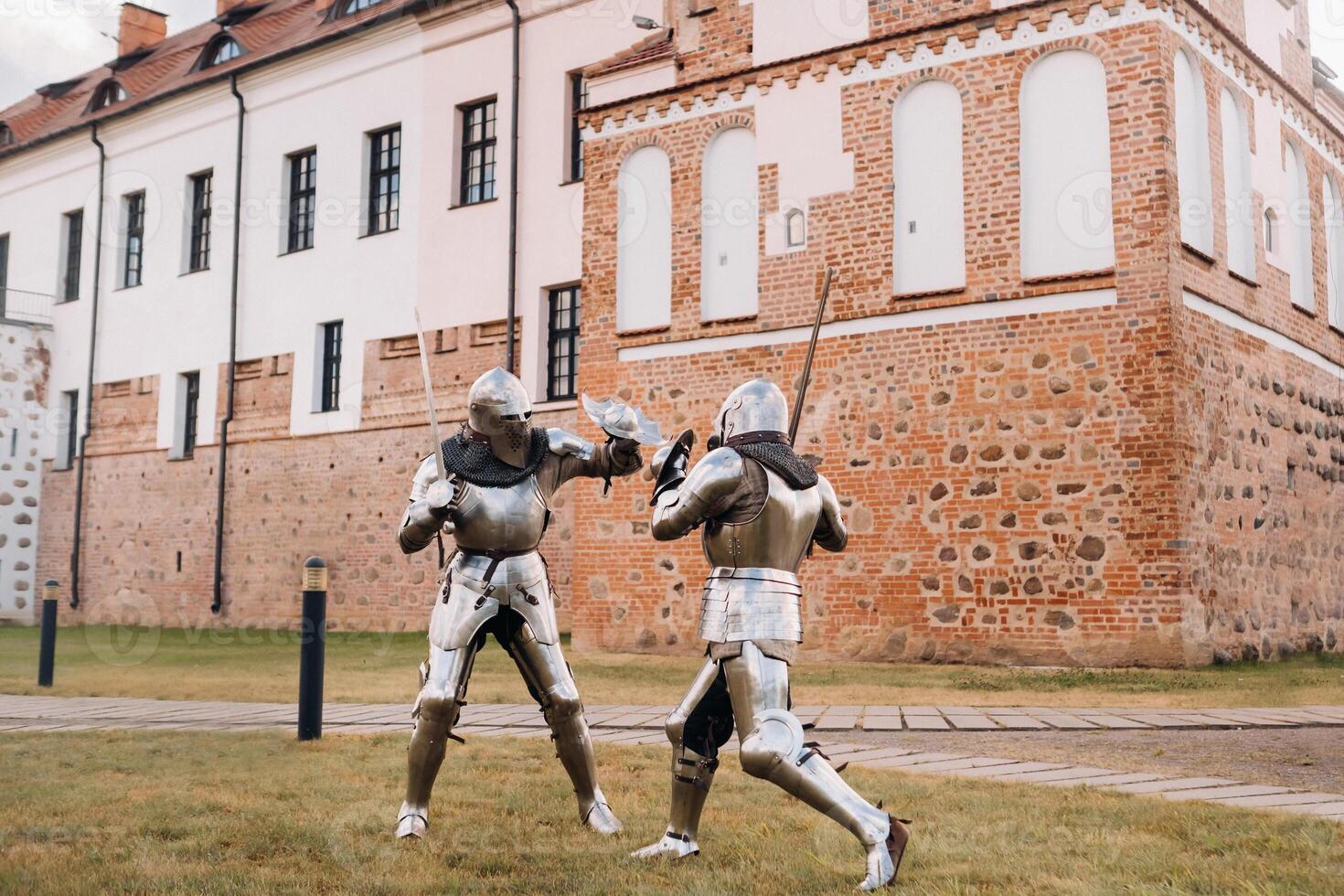 two knights in ancient metal armor are standing at the stone wall of the castle fighting with swords photo