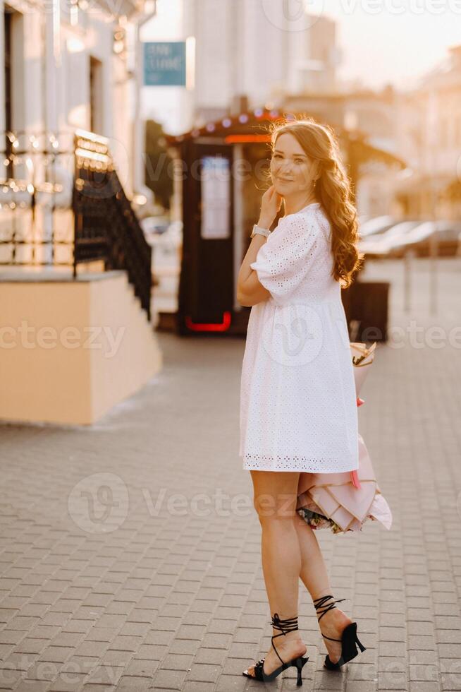 A happy woman in a white dress at sunset with a bouquet of flowers in the city photo