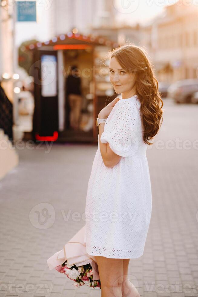 A happy woman in a white dress at sunset with a bouquet of flowers in the city photo