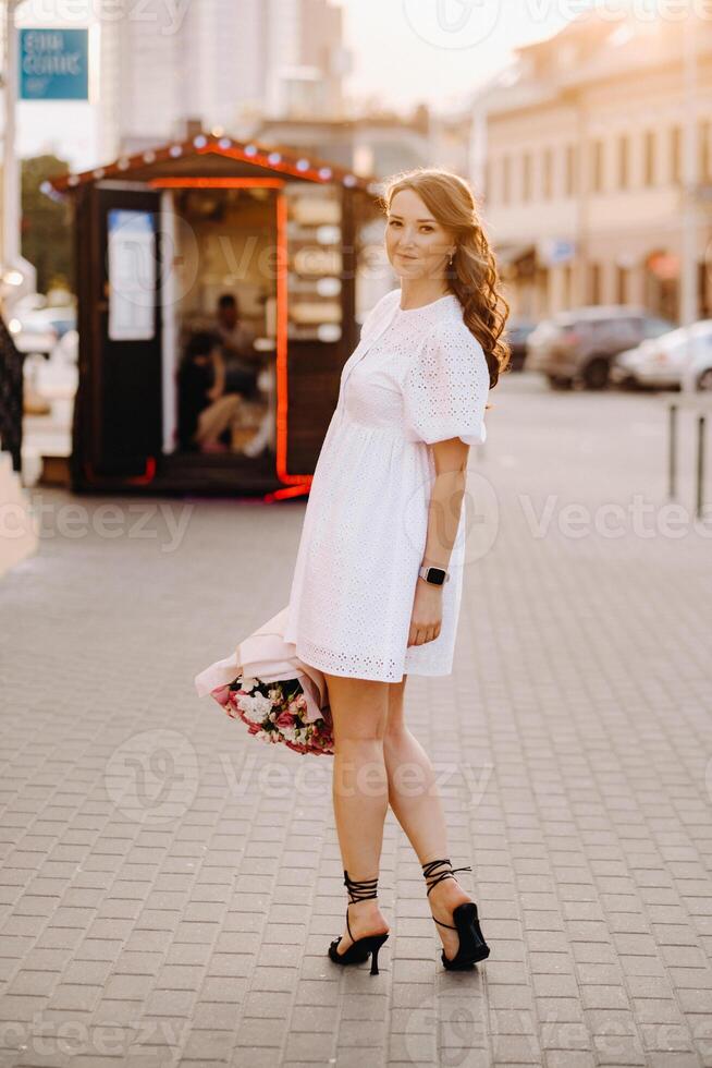 A happy woman in a white dress at sunset with a bouquet of flowers in the city photo