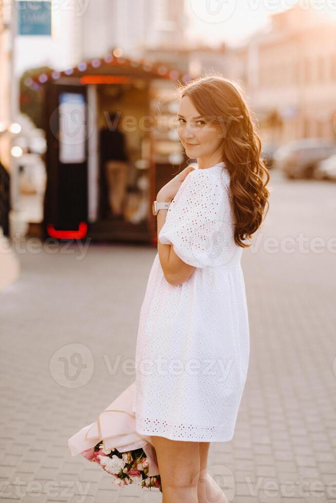 A happy woman in a white dress at sunset with a bouquet of flowers in the city photo