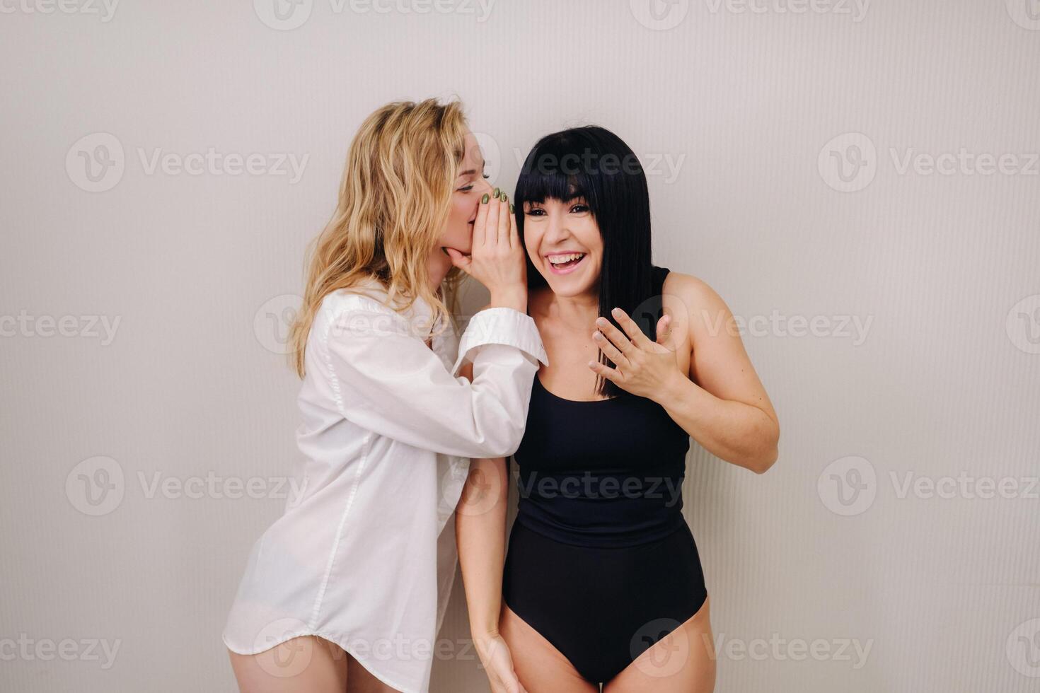 An excited woman in a black swimsuit listens to a friend in a shirt whispering in her ear on a light background photo