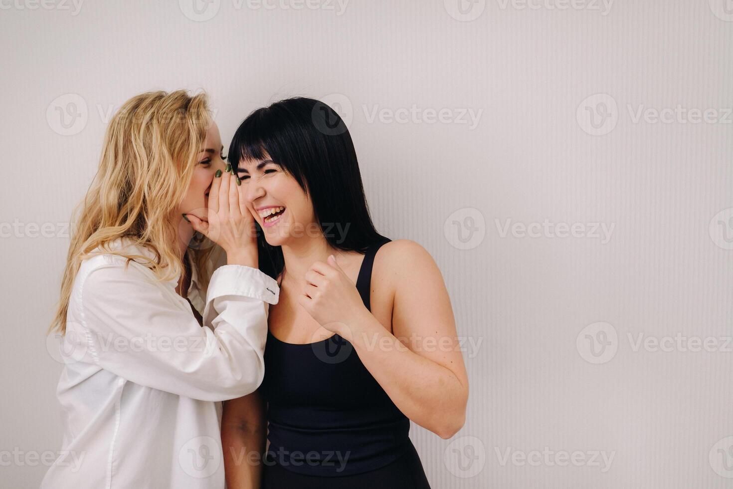 An excited woman in a black swimsuit listens to a friend in a shirt whispering in her ear on a light background photo