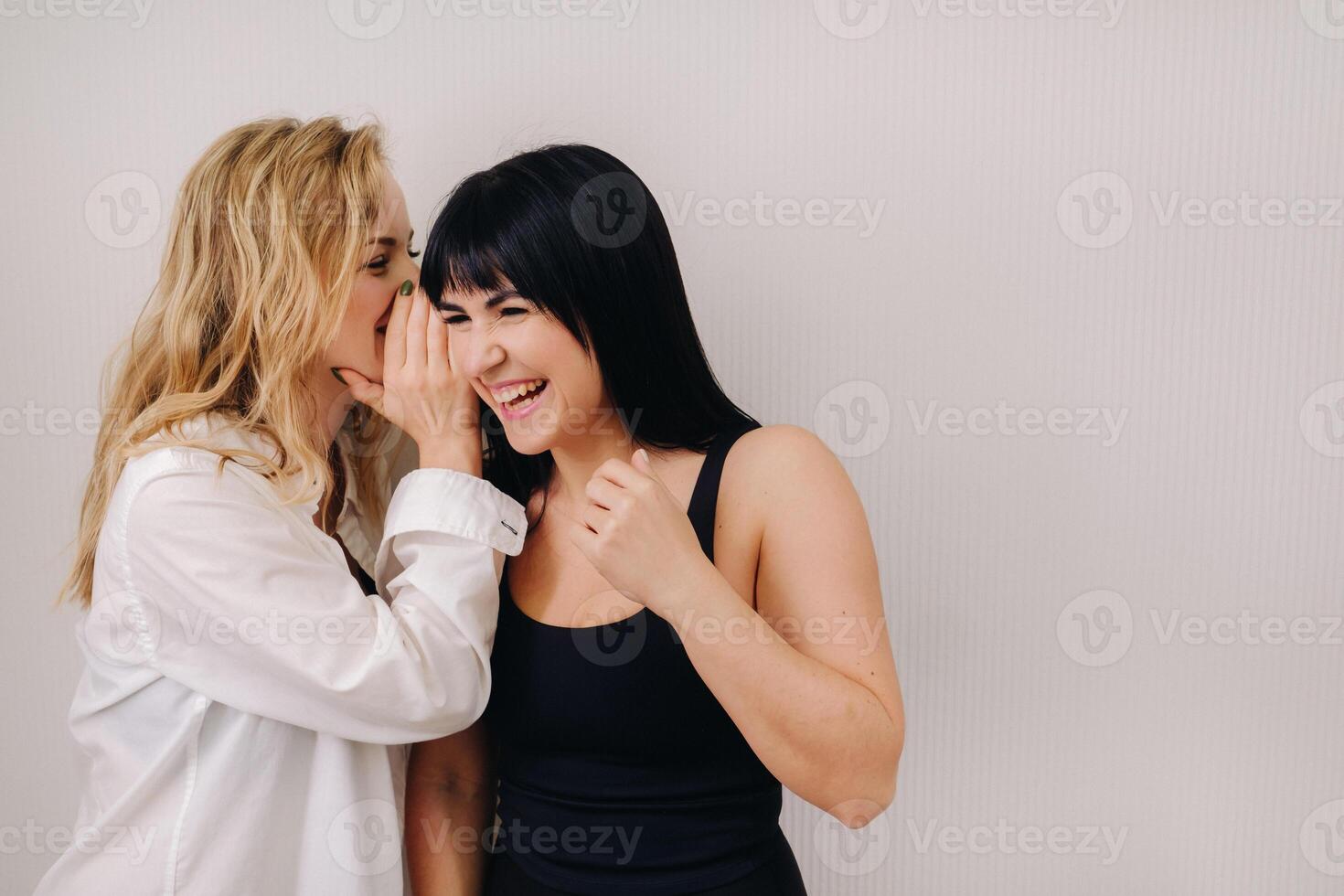 An excited woman in a black swimsuit listens to a friend in a shirt whispering in her ear on a light background photo