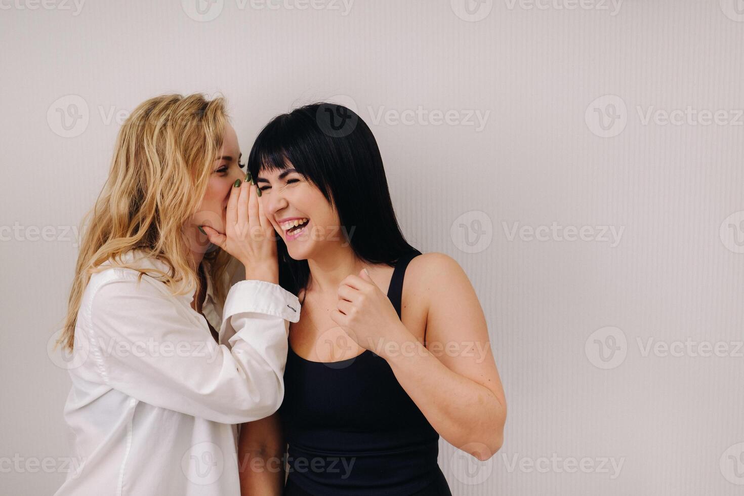 An excited woman in a black swimsuit listens to a friend in a shirt whispering in her ear on a light background photo
