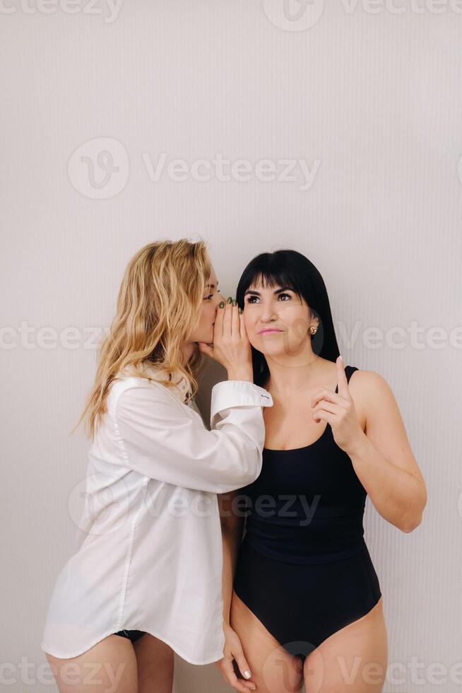 An excited woman in a black swimsuit listens to a friend in a shirt whispering in her ear on a light background photo