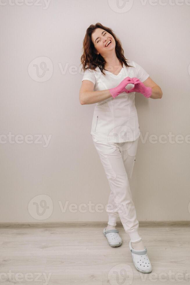 A smiling doctor in pink gloves and a white coat looks into the camera and makes a heart with his hands on a light background photo