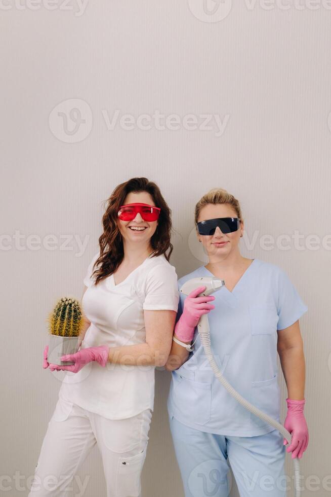 Two cosmetologists in protective glasses are holding a laser depilation device and a cactus in their hands photo