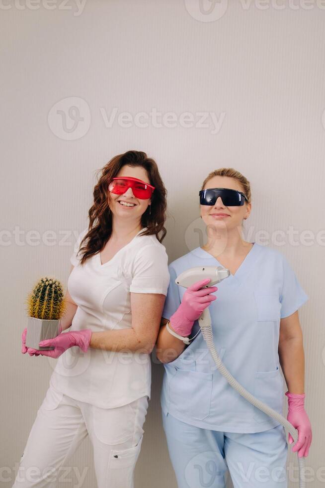 Two cosmetologists in protective glasses are holding a laser depilation device and a cactus in their hands photo