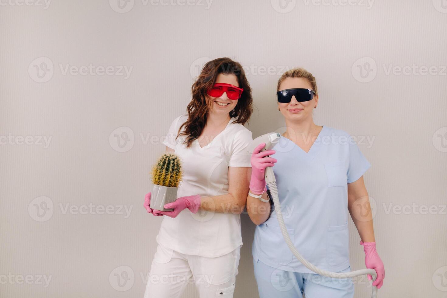 Two cosmetologists in protective glasses are holding a laser depilation device and a cactus in their hands photo