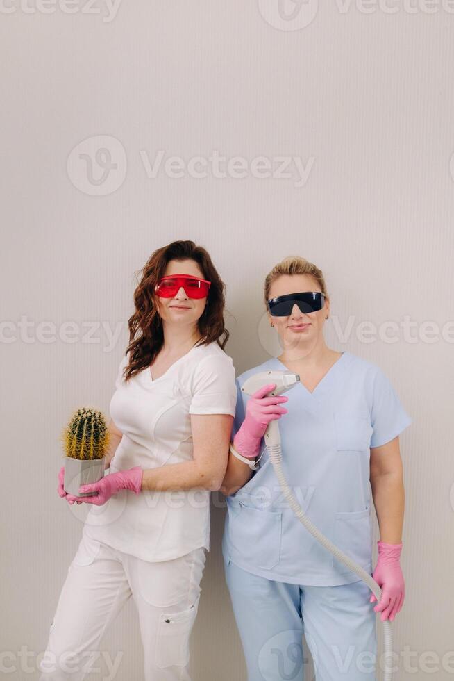 Two cosmetologists in protective glasses are holding a laser depilation device and a cactus in their hands photo