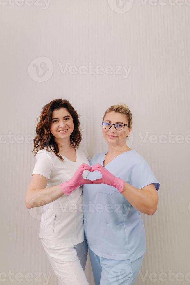 Portrait of two qualified cosmetologists-women, confidently smiling and making a heart with their hands in a medical center photo