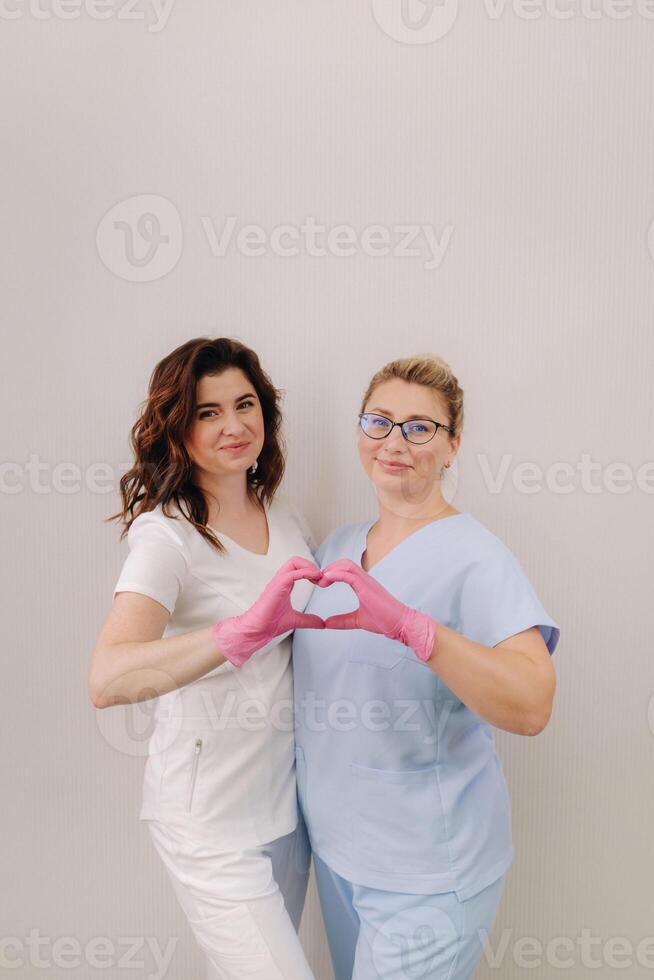 Portrait of two qualified cosmetologists-women, confidently smiling and making a heart with their hands in a medical center photo