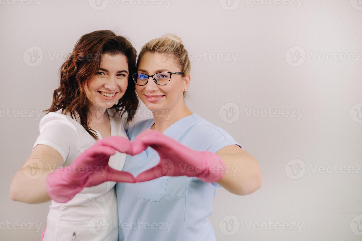 Portrait of two qualified cosmetologists-women, confidently smiling and making a heart with their hands in a medical center photo