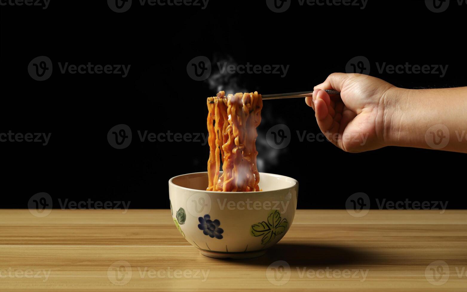 Person holding chopsticks noodles with steam and smoke on black background, Asian noodle food concept, ramen noodles hot and spicy served in a bowl on rustic wooden table, Chinese style. photo