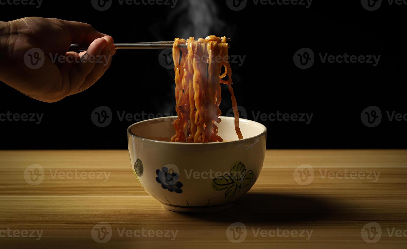 Person holding chopsticks noodles with steam and smoke on black background, Asian noodle food concept, ramen noodles hot and spicy served in a bowl on rustic wooden table, Chinese style. photo