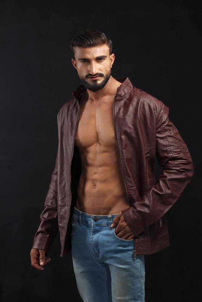 portrait of a young pakistani gym fit male model showing his build physique bicep triceps arms shoulder wings along with brown jacket photo