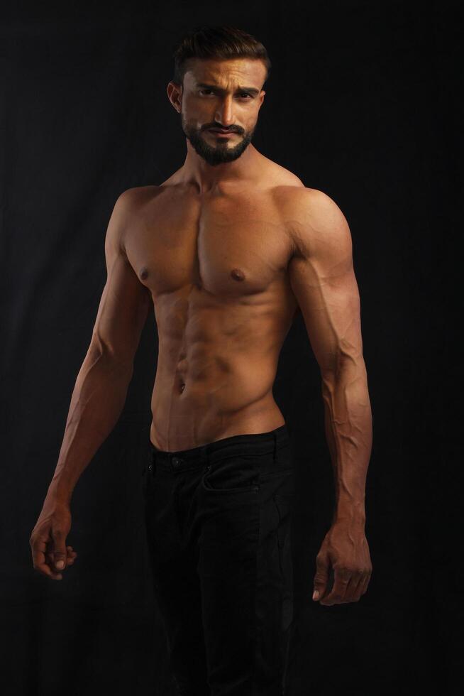 portrait of a young pakistani gym fit shirtless male model showing his build physique bicep triceps arms shoulder wings against black background photo
