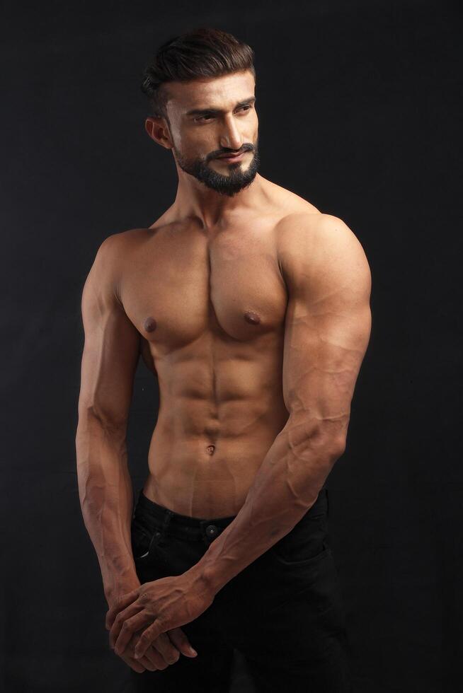 portrait of a young pakistani gym fit shirtless male model showing his build physique bicep triceps arms shoulder wings against black background photo