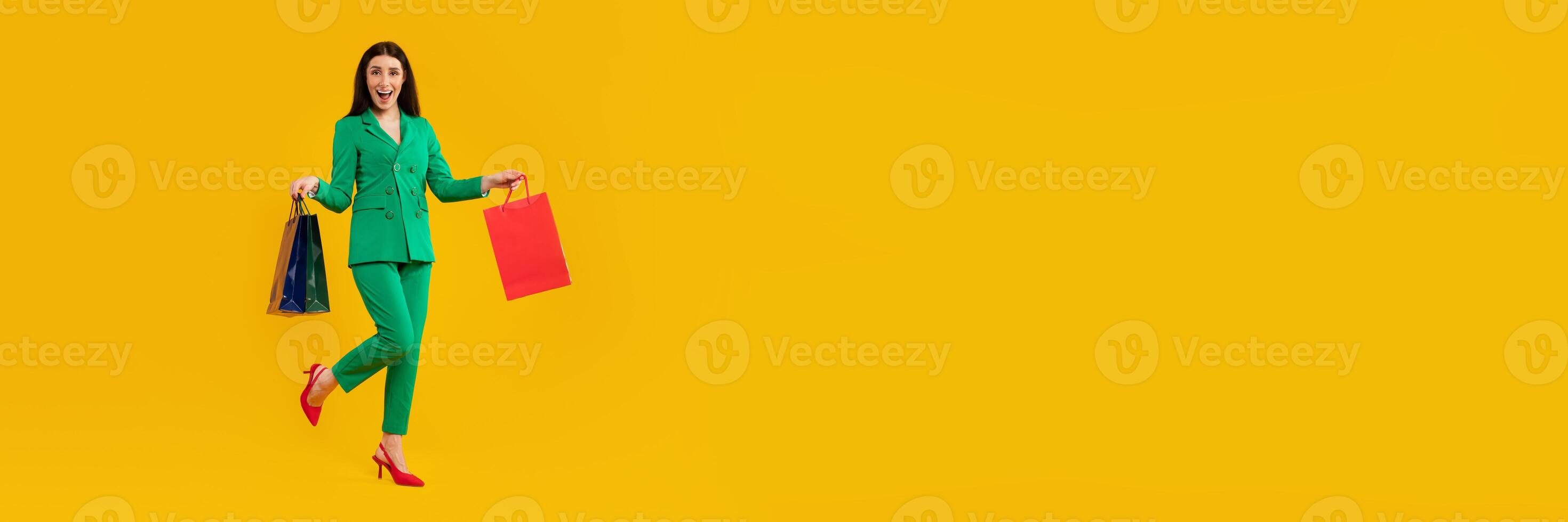 A cheerful woman in a bright green suit holds shopping bags while celebrating a Black Friday sale. The vibrant yellow background enhances the upbeat shopping atmosphere, ideal for promotional use. photo