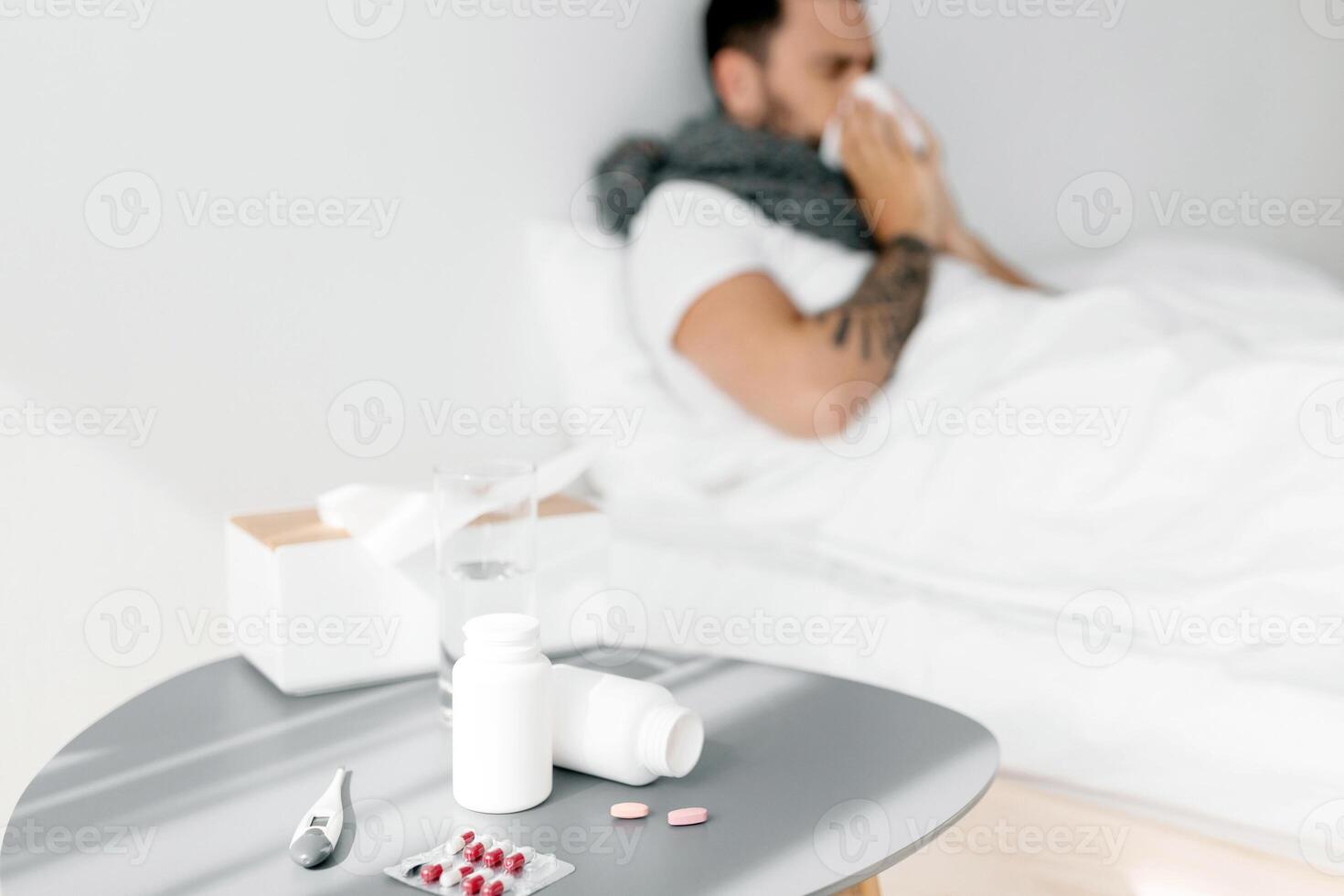 Sick millennial man sneezing in paper tissue, lying under blanket in bed, suffering from seasonal flu and cold. Focus on bed table full of medicine pills photo