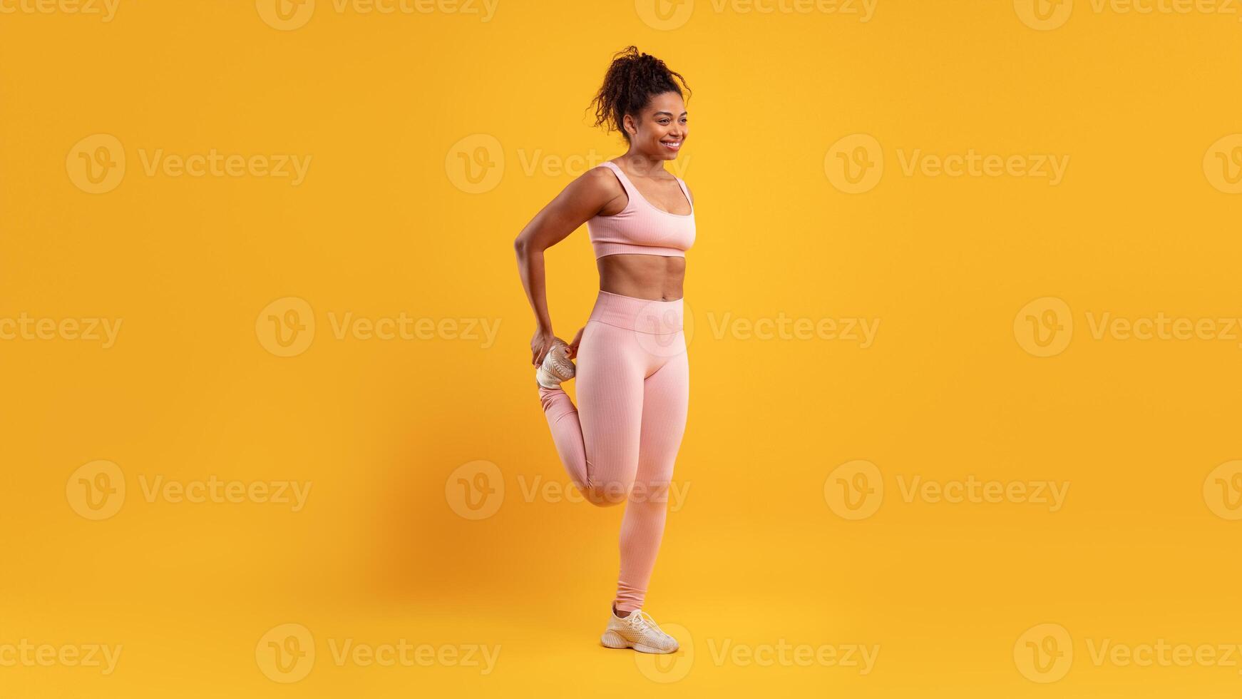 A young woman in pink activewear performs a leg stretch before starting her exercise routine in a vibrant yellow studio. Her focused expression shows dedication to fitness. photo