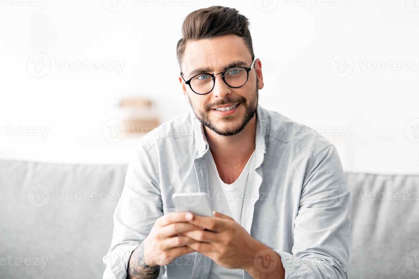 Positive young guy with glasses sitting on couch, using smartphone and smiling to camera, chatting with friends and chilling at home, copy space photo