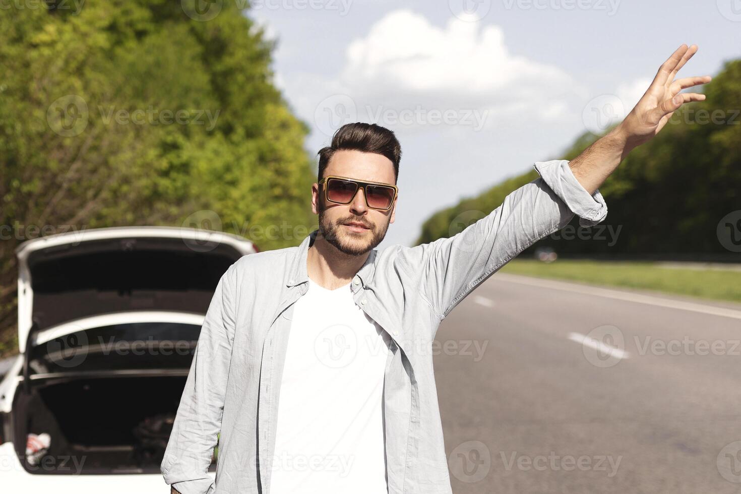 Young Caucasian man trying to wave down car, asking for help, his friends trying to fix broken vehicle, having flat tire on road. Millennial guy hitchhiking on highway, needing emergency assistance photo