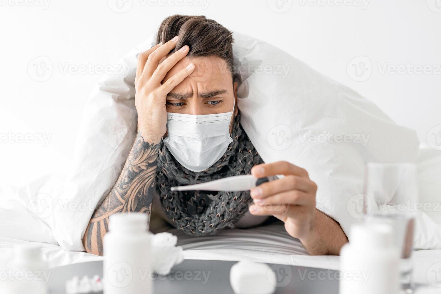 Shocked sick guy in mask looking at thermometer with high temperature result, lying under blanket in bed at home. Covid-19 coronavirus disease, seasonal flu and cold, illness fatigue photo