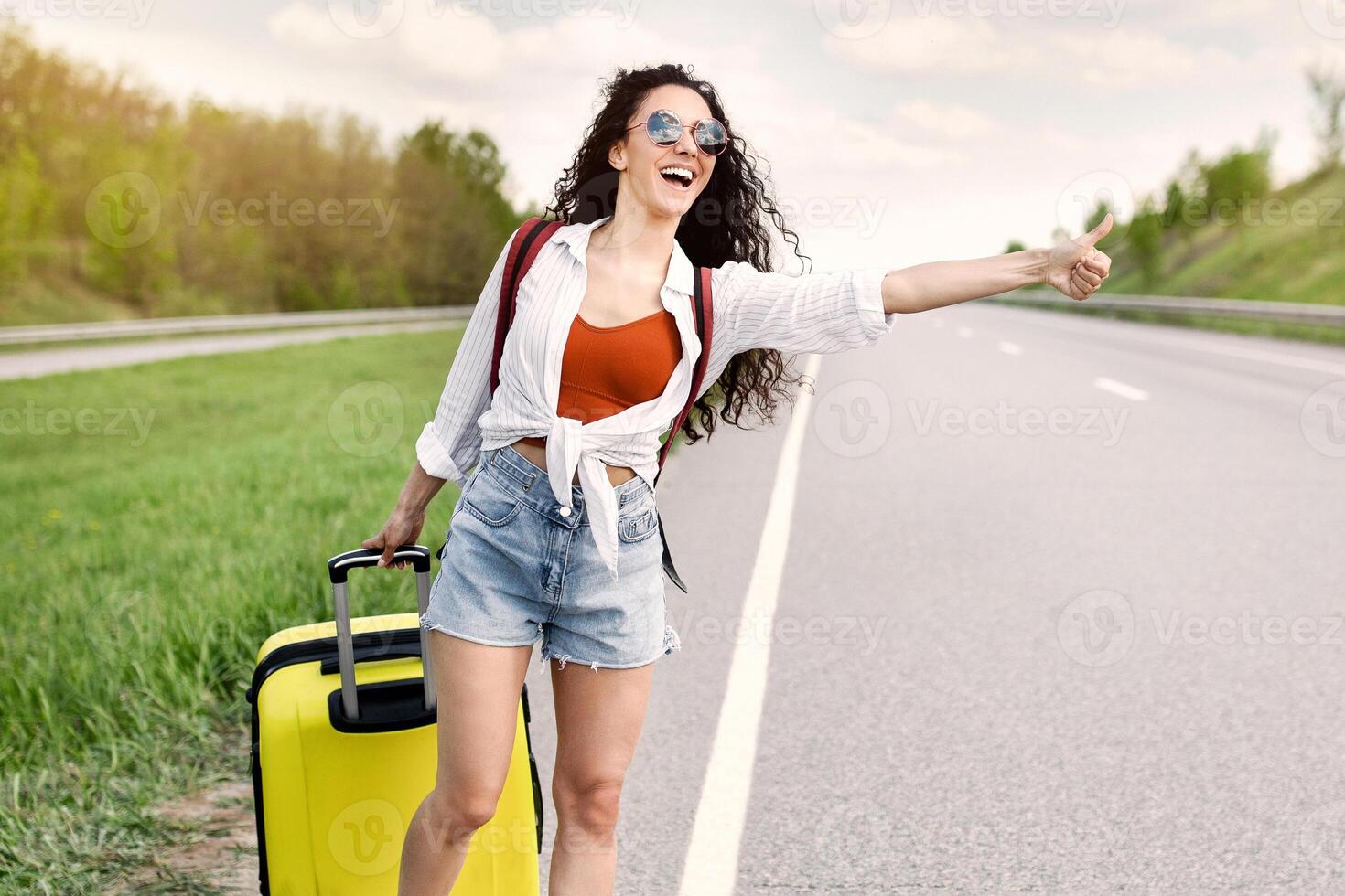 Joyful millennial lady thumbing her ride, catching car with suitcase on highway, hitchhiking in countryside, copy space. Summertime adventre, autostop journey concept photo