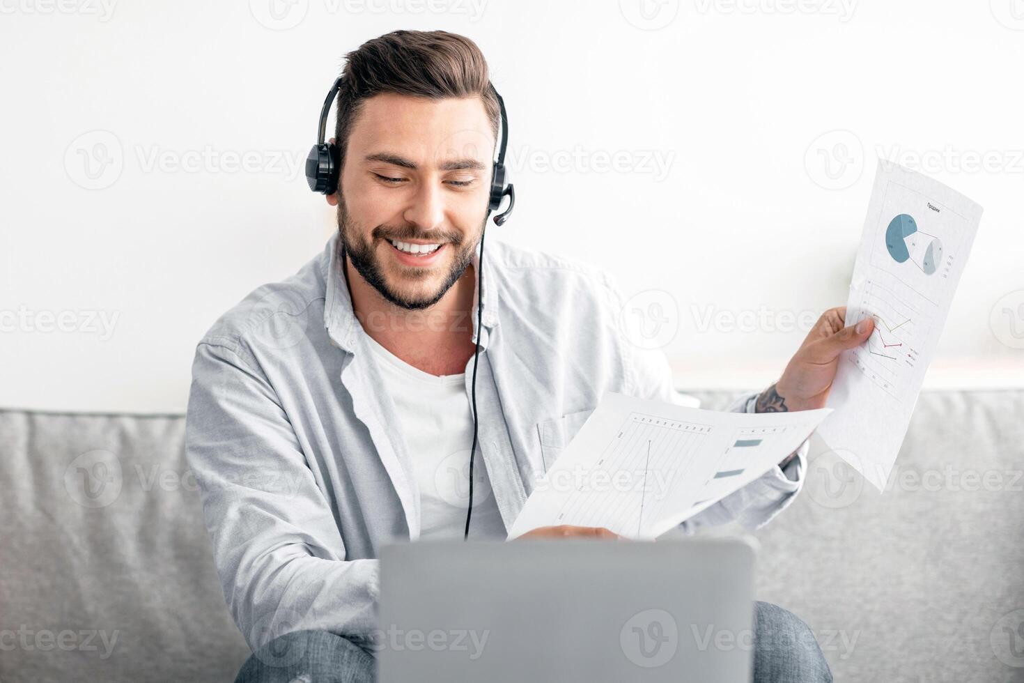 Virtual meeting. Happy man having call, showing documents with diagrams to laptop camera, discussing financial reports, wearing headset while sitting at home interior photo