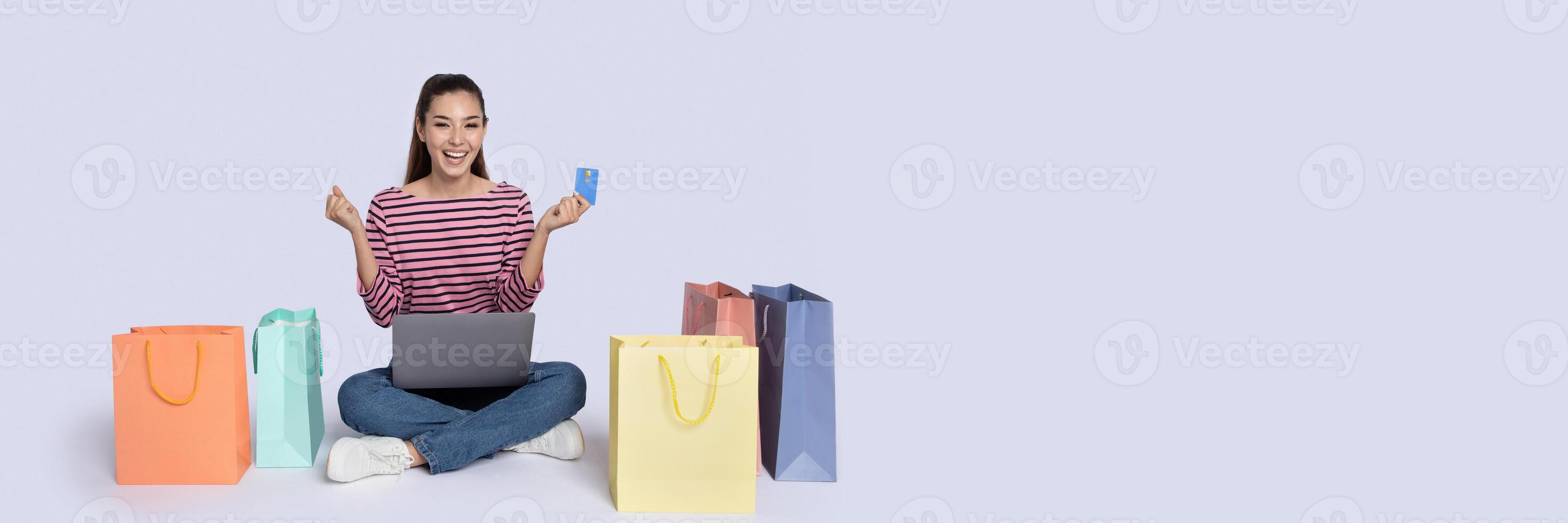 A happy shopper sits on the floor surrounded by colorful shopping bags while holding a credit card and a laptop. She enjoys the thrill of online shopping for great Black Friday deals, copy space photo