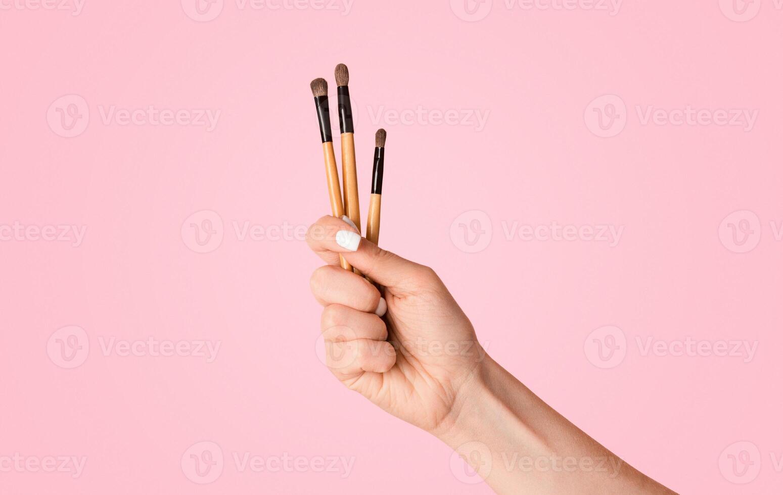 Closeup view of beautician's hand showing lipstick or eyeshadow brushes on pink background photo