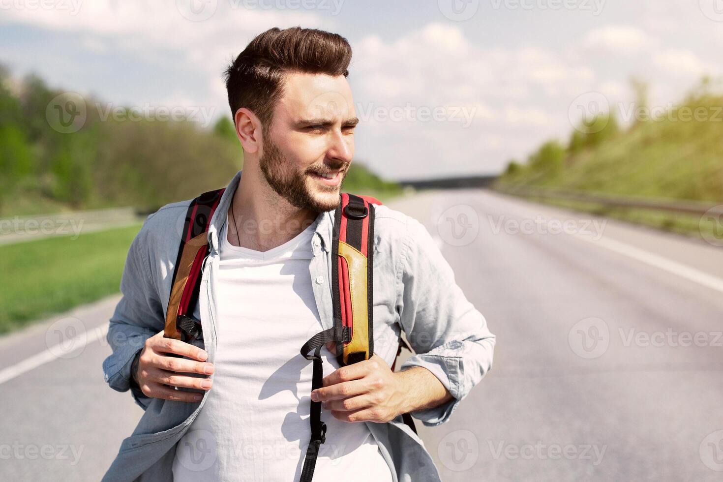 Handsome young man with backpack walking along highway, traveling by autostop, hitchhiking in countryside. Cheerful Caucasian guy having summertime adventure, enjoying holidays photo