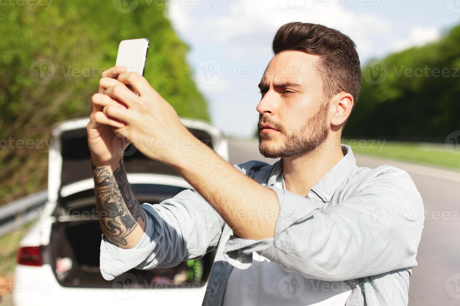 Upset young guy standing on road next to broken car, looking for mobile network signal, trying to call roadside assistance, having no connection, his friends trying to fix flat tire, copy space photo