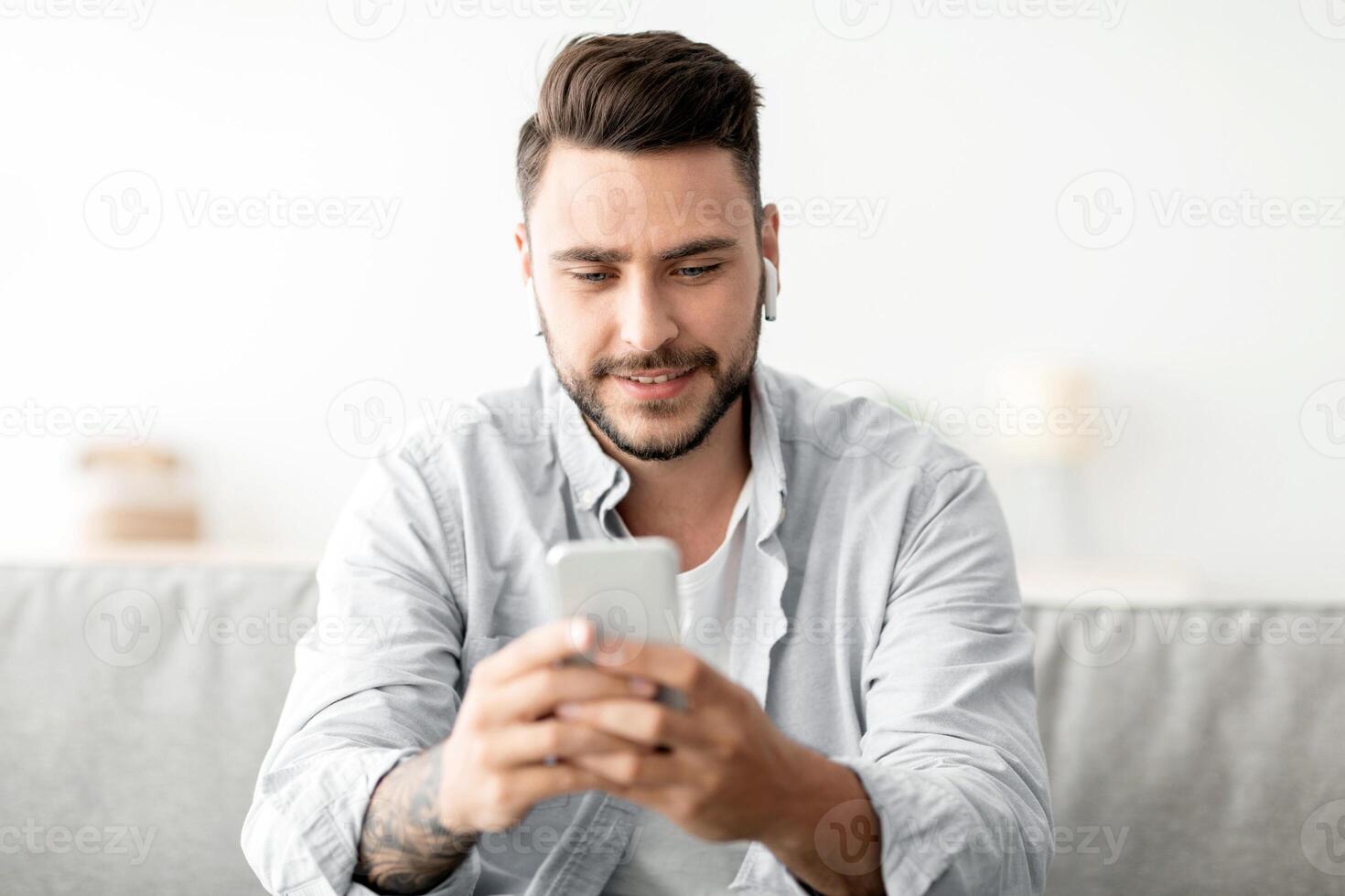 Young man with earbuds using smartphone, browsing internet and listening to music online, having call while sitting on couch at home. Cellphone application concept photo