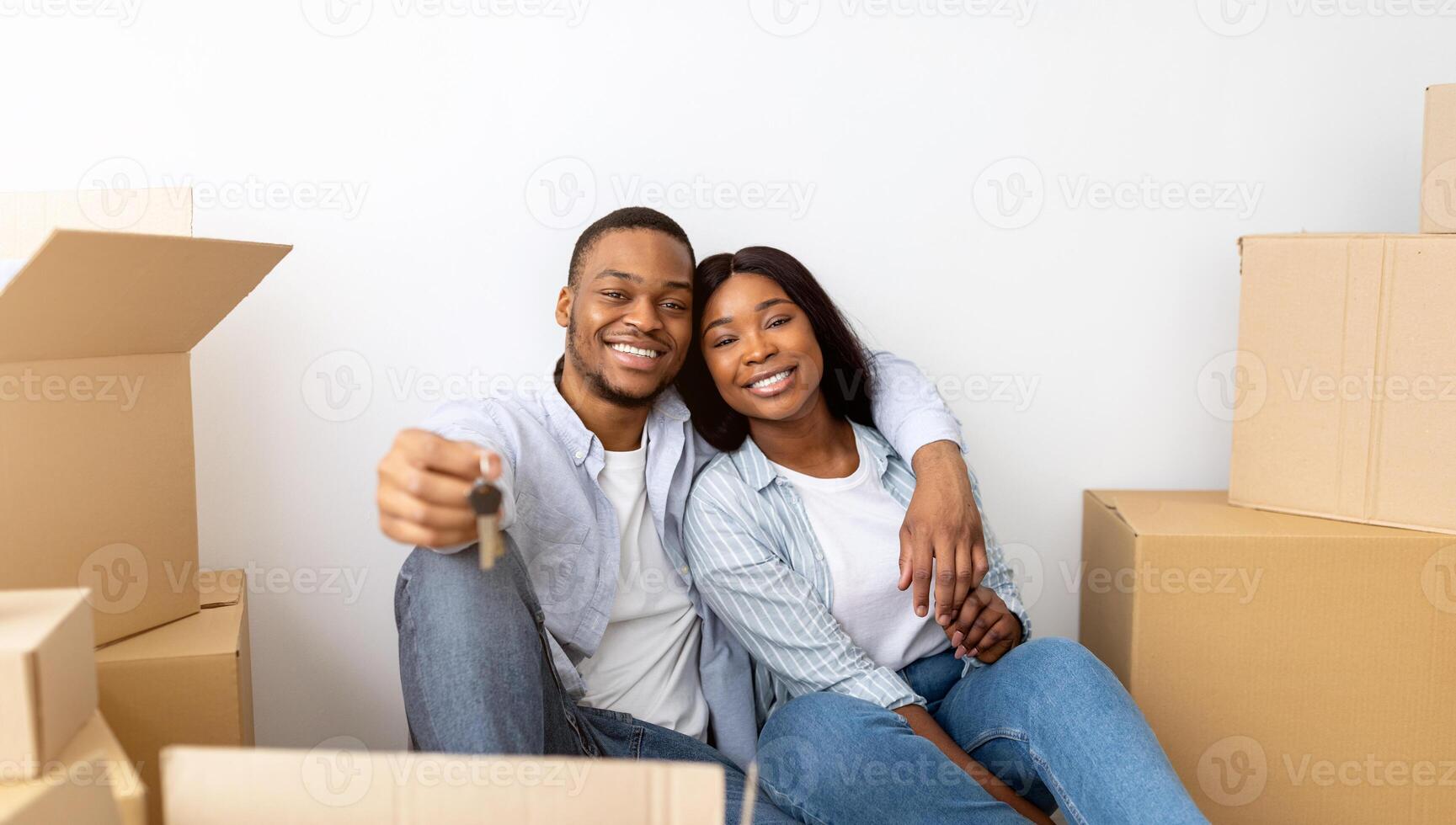 House ownership. Loving black couple holding keys, sitting among cardboard boxes and hugging after moving in new apartment. Insurance, real estate, mortgage concept photo
