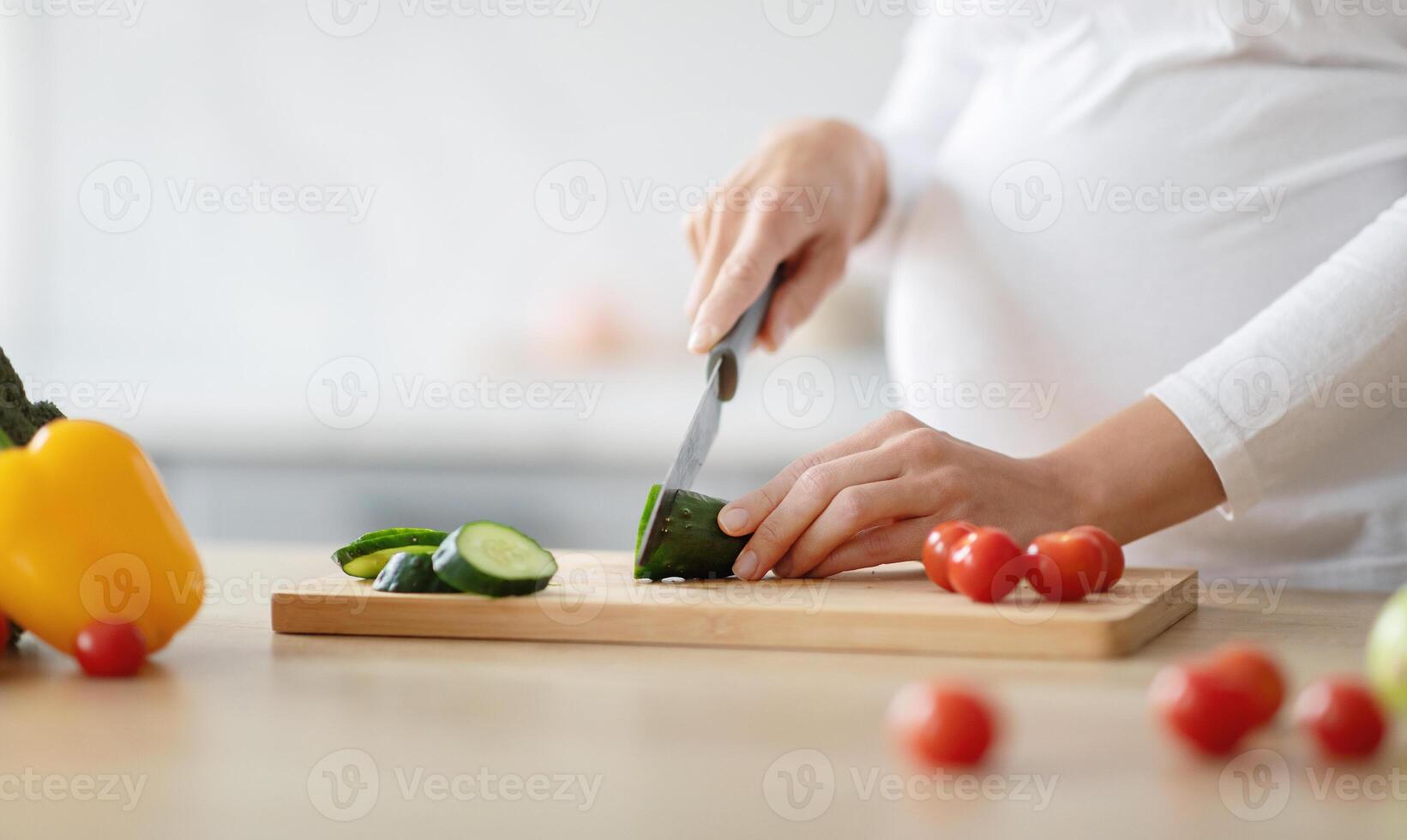 A pregnant woman is using a knife to slice cucumber on a wooden cutting board. Bright tomatoes and a yellow pepper are scattered around, indicating healthy meal preparation. photo