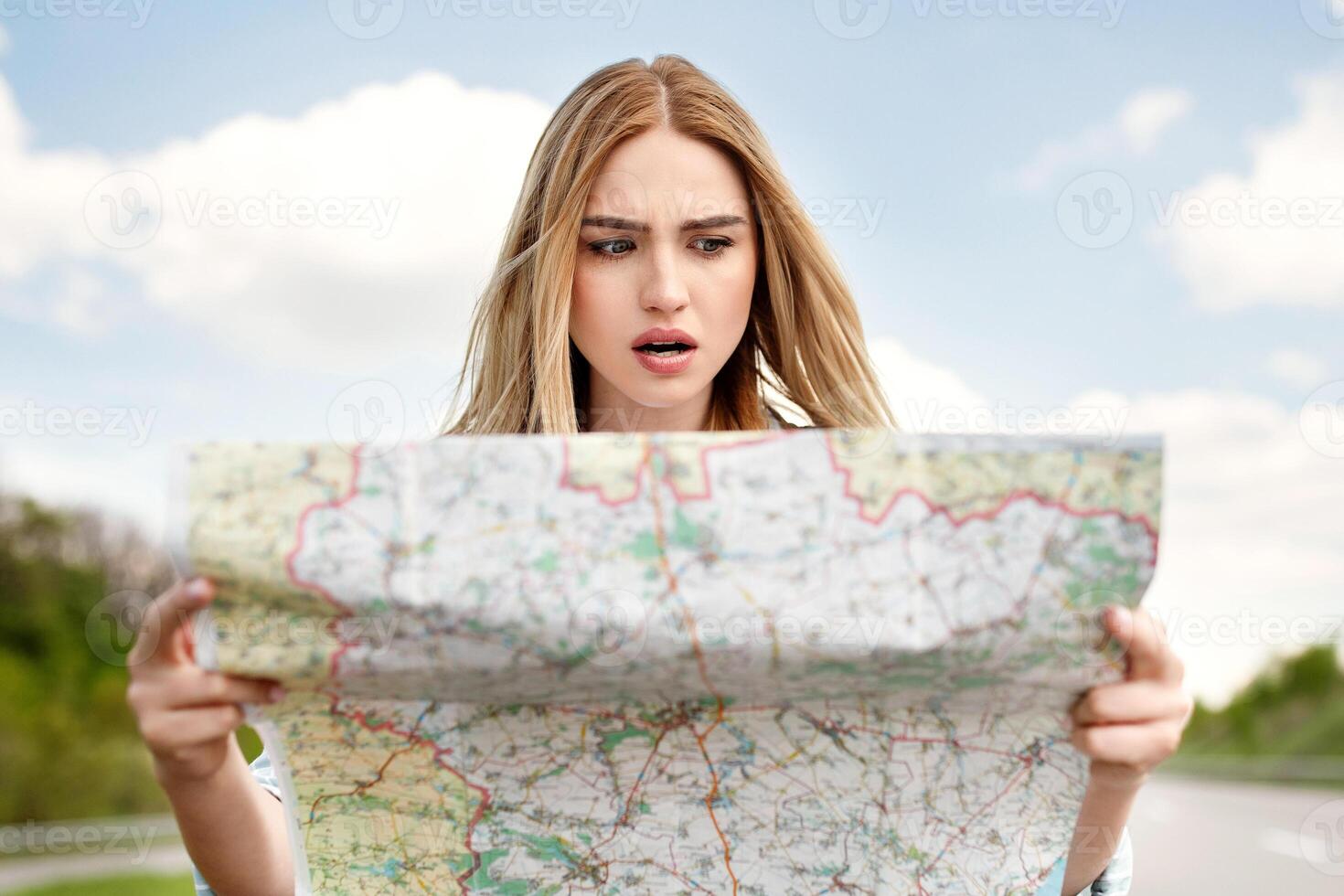 Shocked young blonde woman hitchhiking on road, checking map, feeling lost and confused, traveling alone by autostop. Millennial Caucasian lady standing on highway, cannot find her destination photo